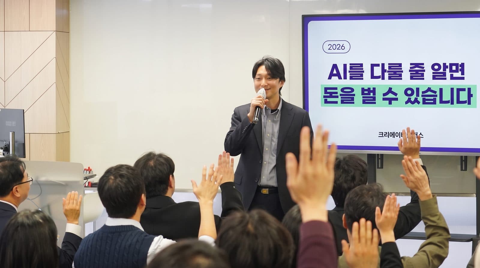 강연장에서 마이크를 든 강사가 이야기하고, 수강생들이 손을 들어 참여하는 모습. 뒤 화면에 'AI를 다룰 줄 알면 돈을 벌 수 있습니다'라고 적혀 있다